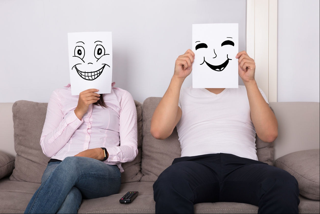Couple Holding Smiling Faces In Front Of Their Faces Sitting On Couch